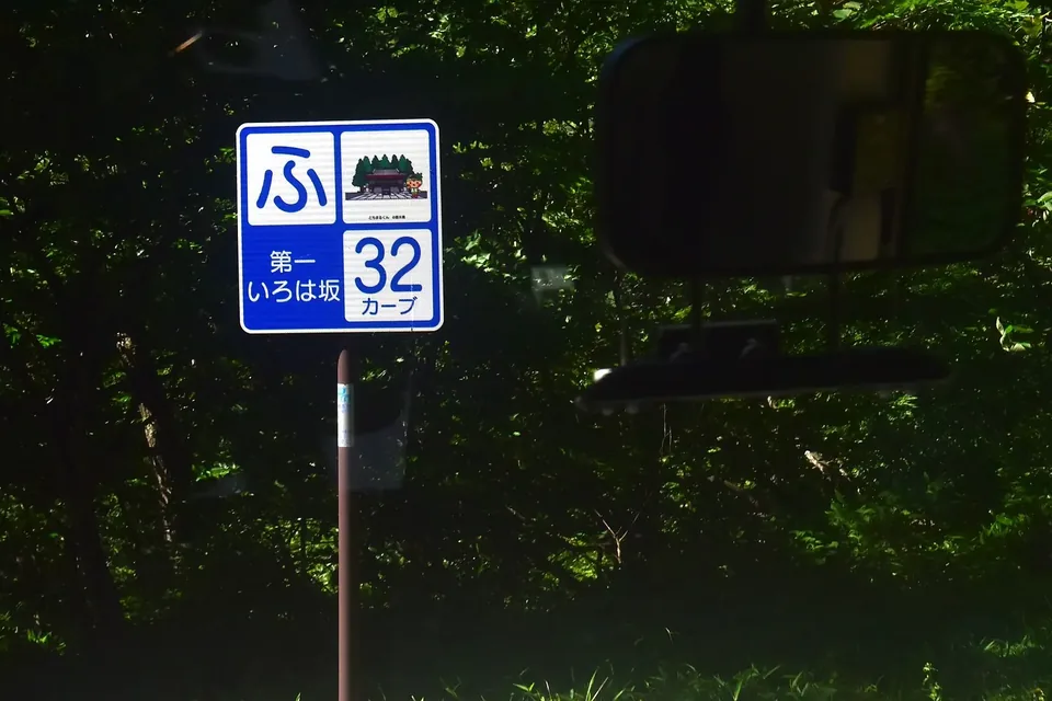 木々に囲まれた道路脇に立つ、ひらがなの「ふ」と「第一いろは坂 32 カーブ」を示す青い標識